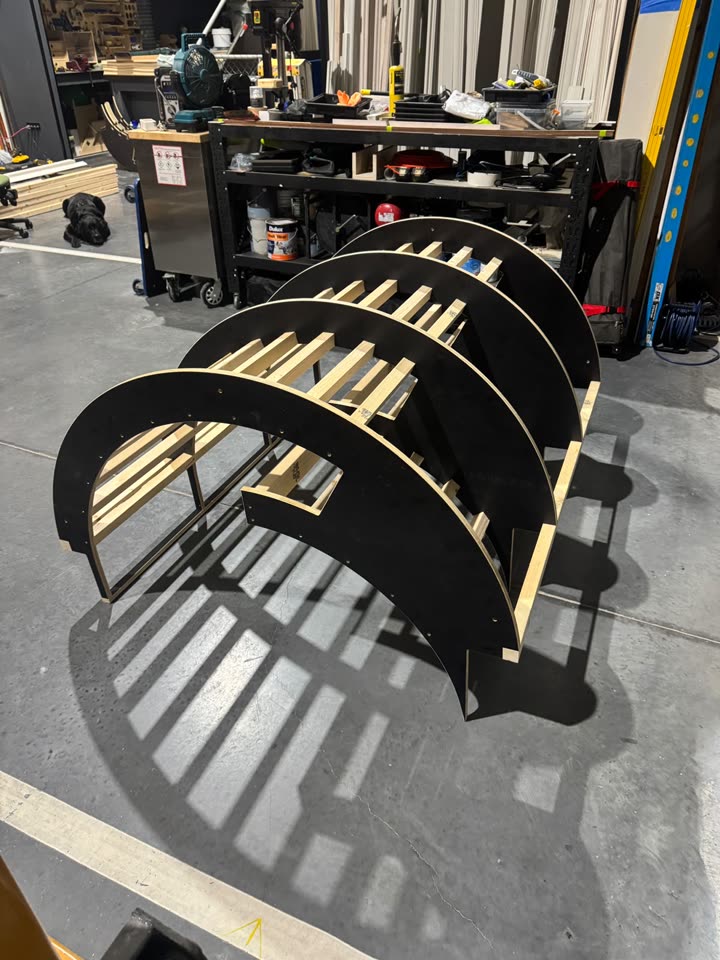 Black formply arch ribs joined by pine battens, standing on the workshop floor during a dry fit.